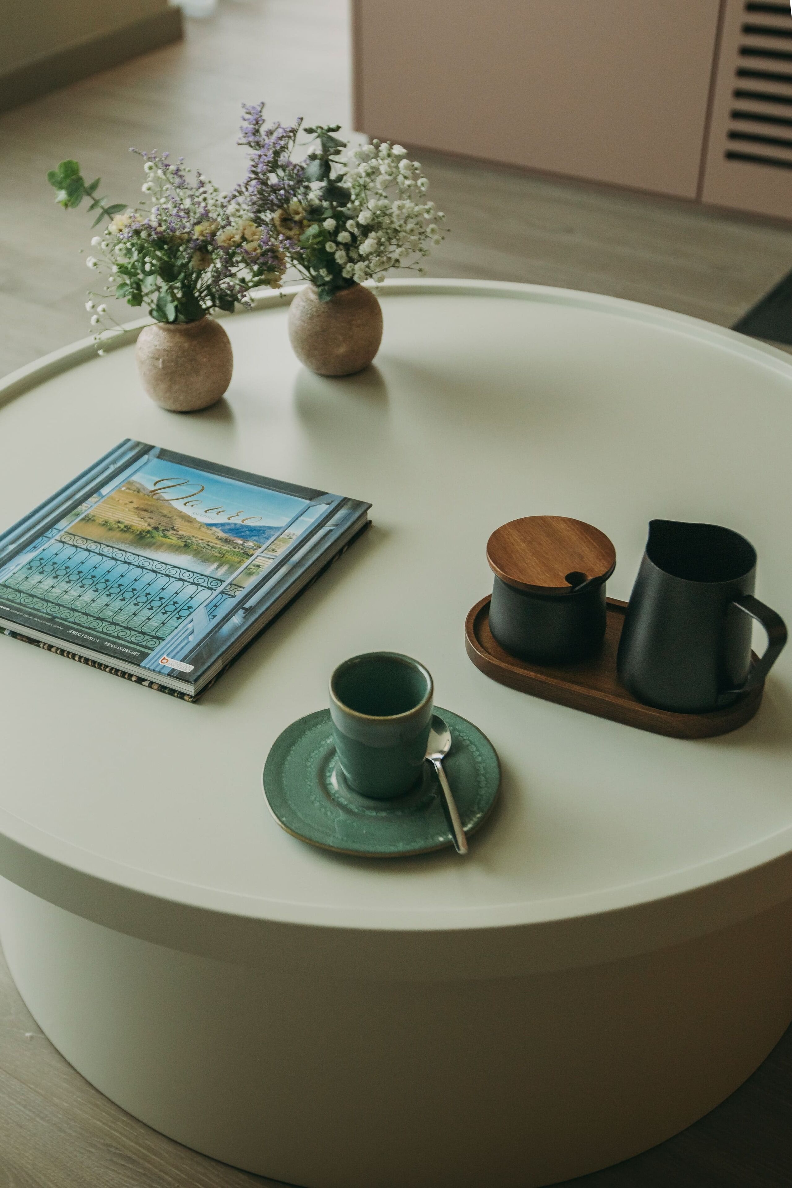 Una mesa redonda blanca con un libro, una taza verde, una bandeja de madera con una mug negra y dos jarrones pequeños con flores.