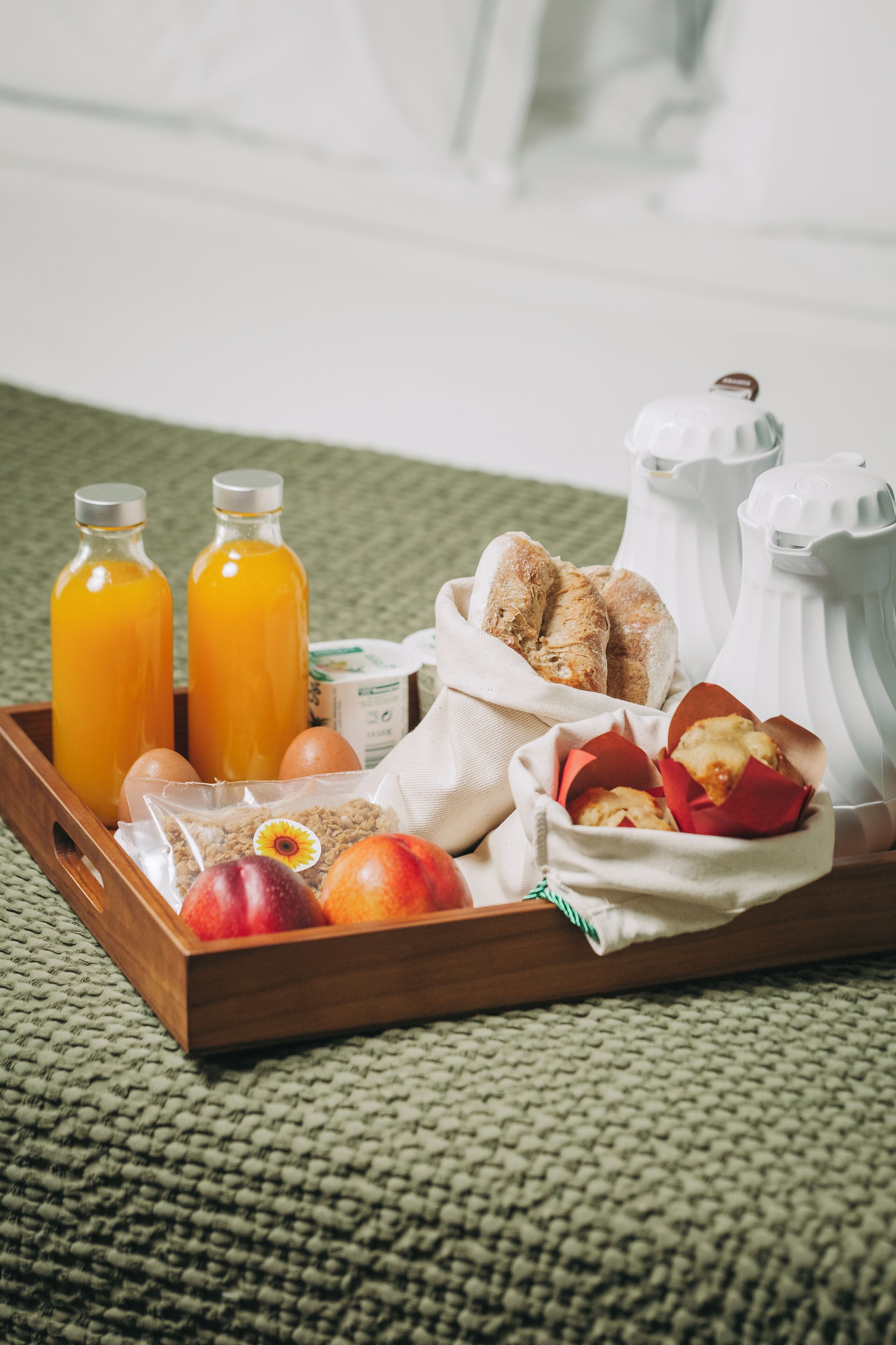 A wooden tray with breakfast items including juice bottles, eggs, bread, and pastries on a green textured surface.