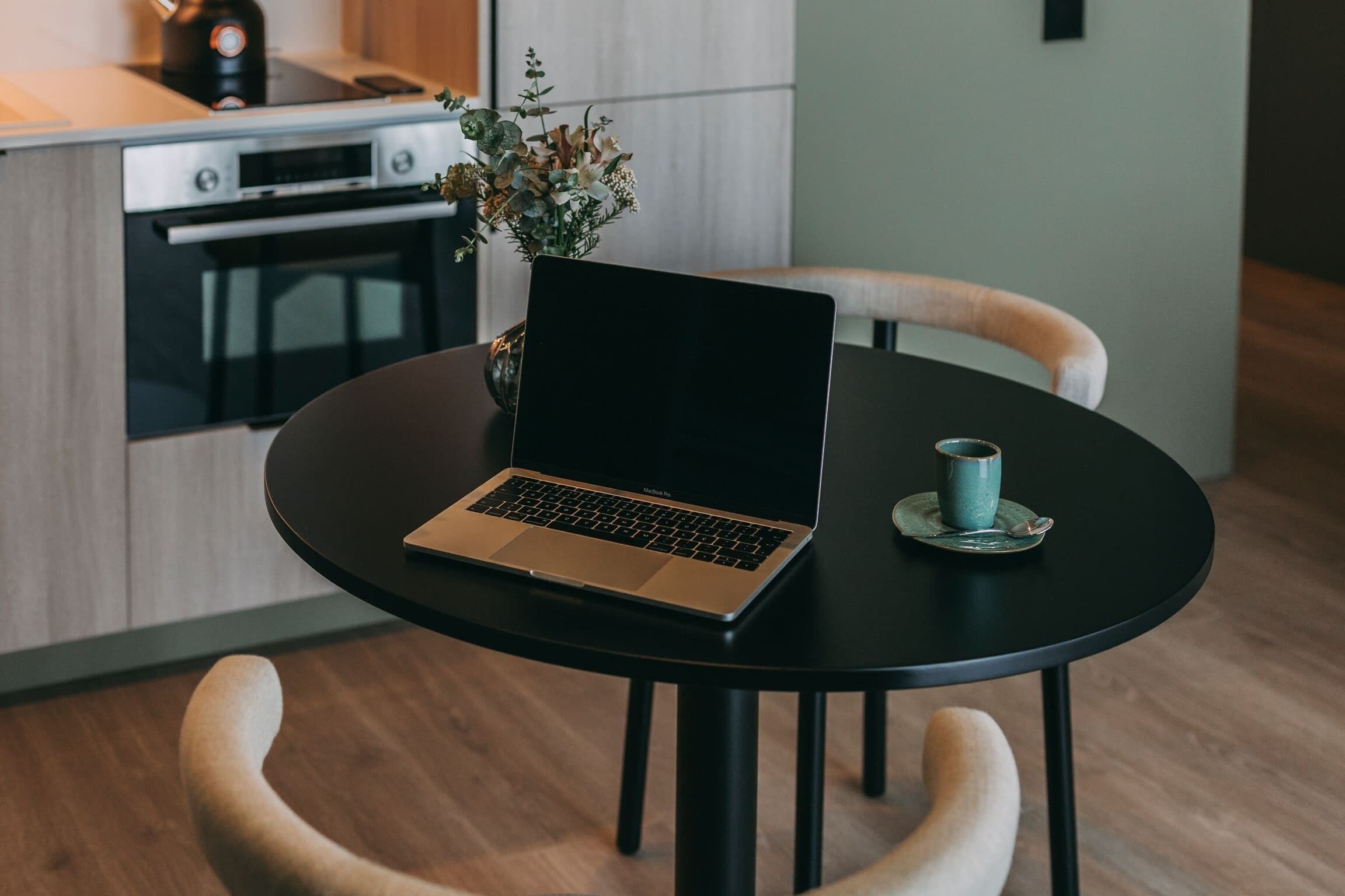 A modern kitchen with a laptop on a round table, a vase of flowers, and a cup on the table.