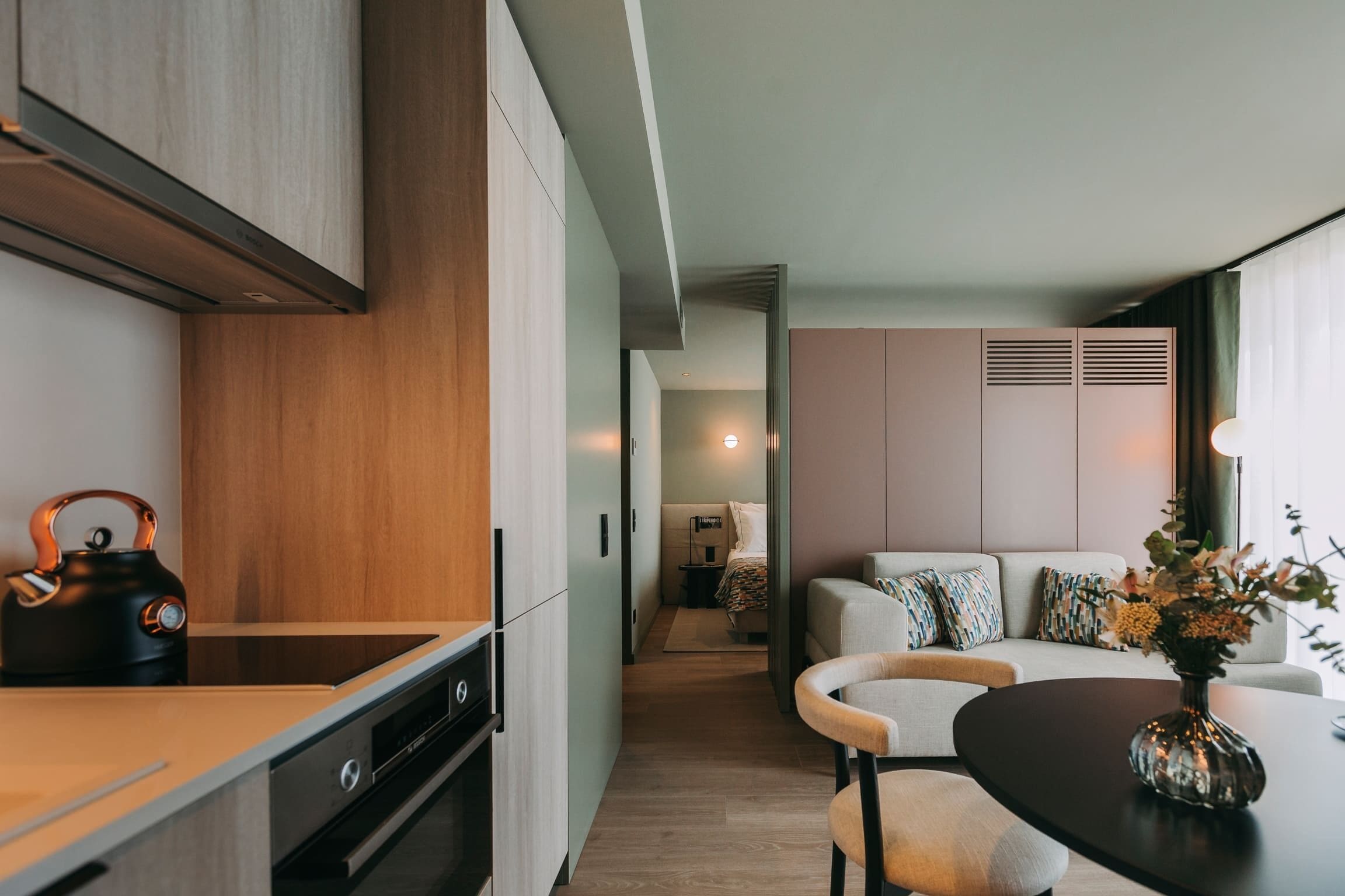 A modern kitchen with a kettle, a dining table with chairs, and a sofa in the adjacent living area.
