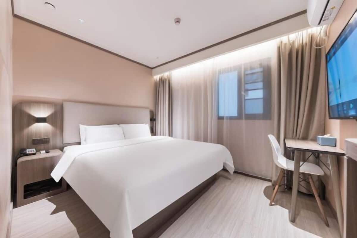 A modern hotel room with a large bed, a desk with a chair, a TV, and a window with curtains.