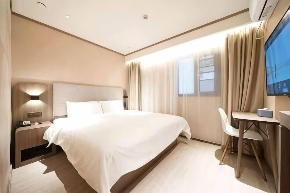 A modern hotel room with a large bed, bedside table, lamp, TV, and a small desk with chair.