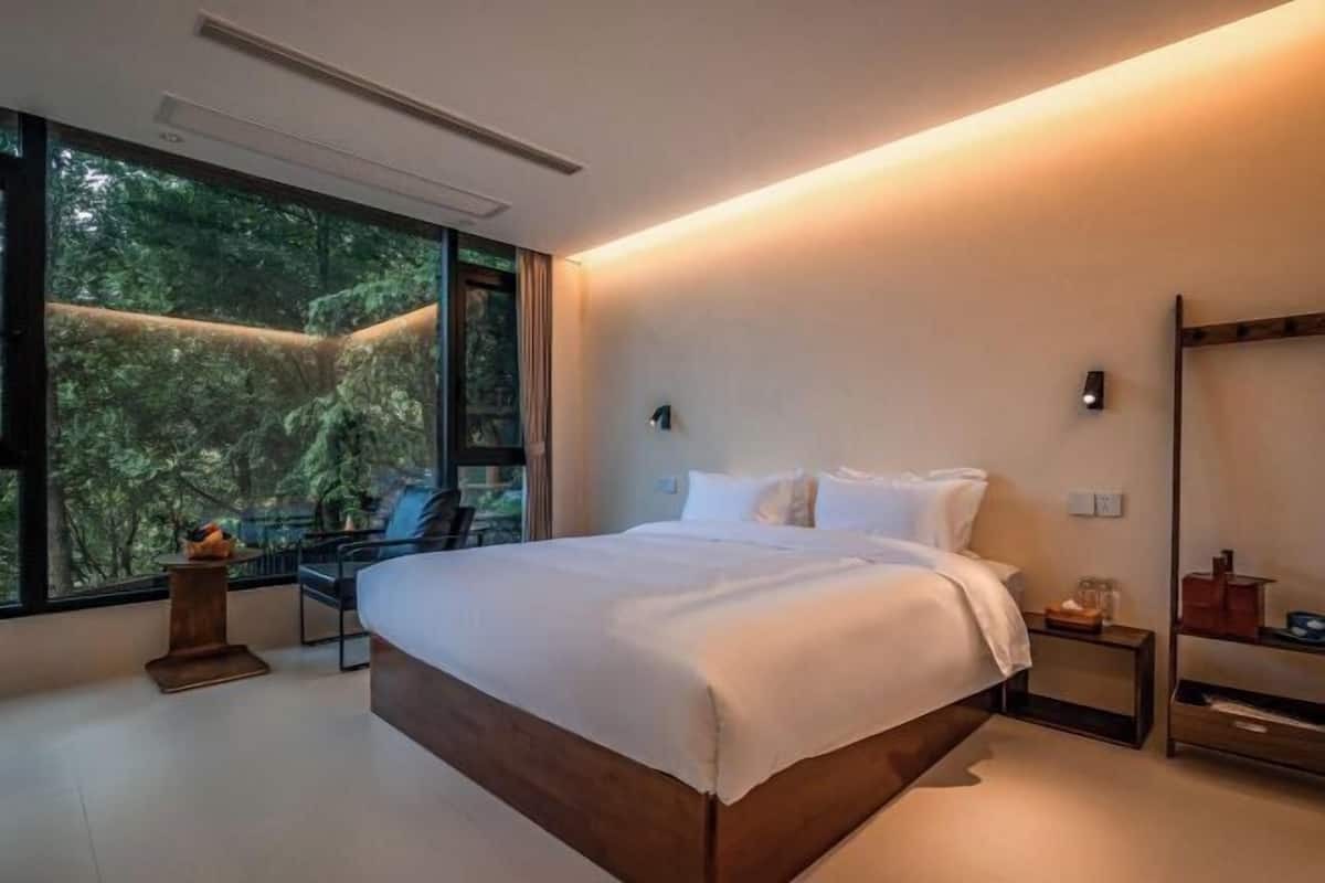 A modern bedroom with a large bed, a desk, and a view of a forest through the window.