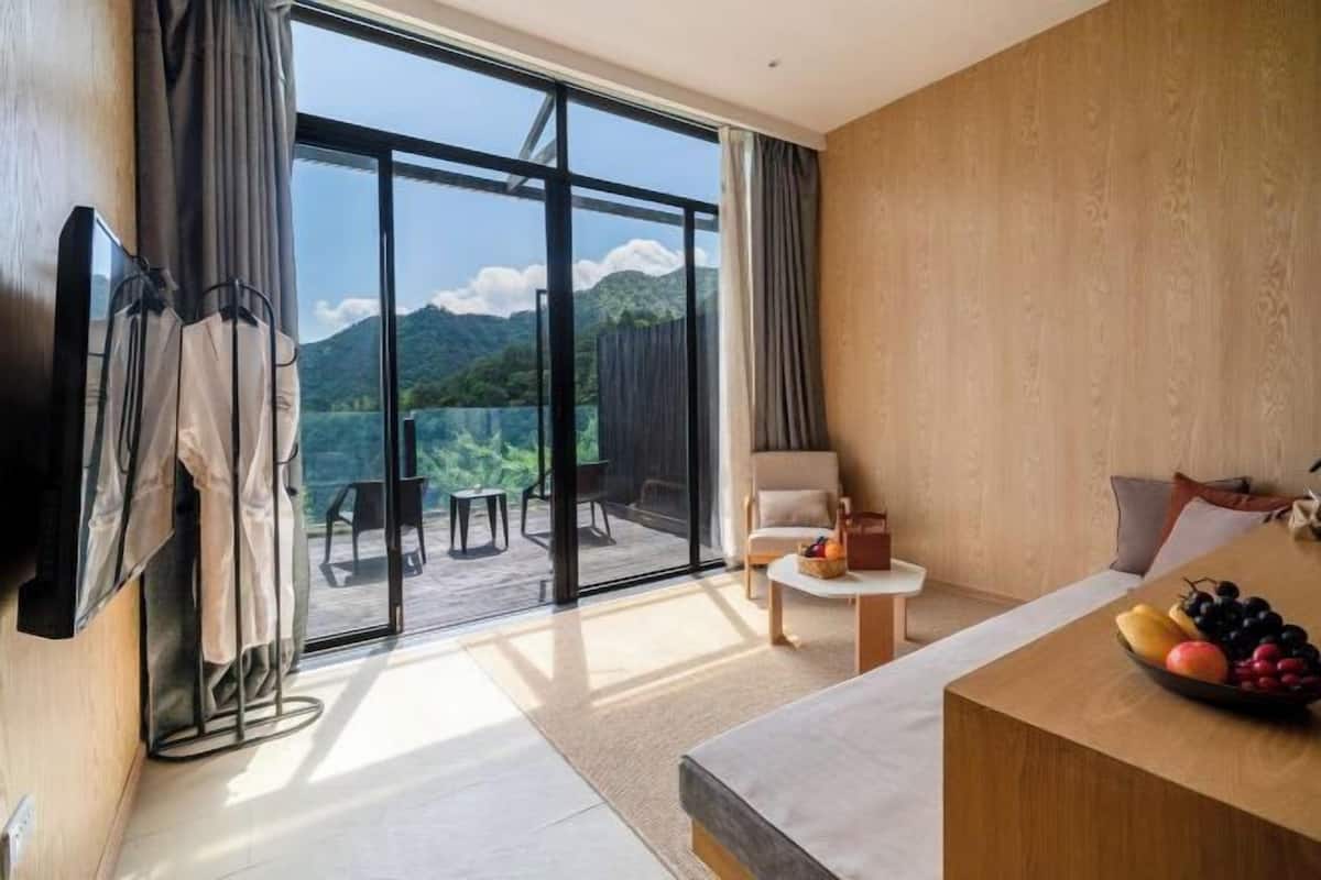 A modern hotel room with a large sliding glass door leading to a balcony, a flat-screen TV mounted on the wall, a seating area with a small table, and a fruit bowl on the side table.