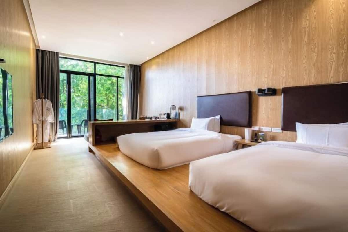 A hotel room with two beds, a wooden bench, a desk, and a large window.