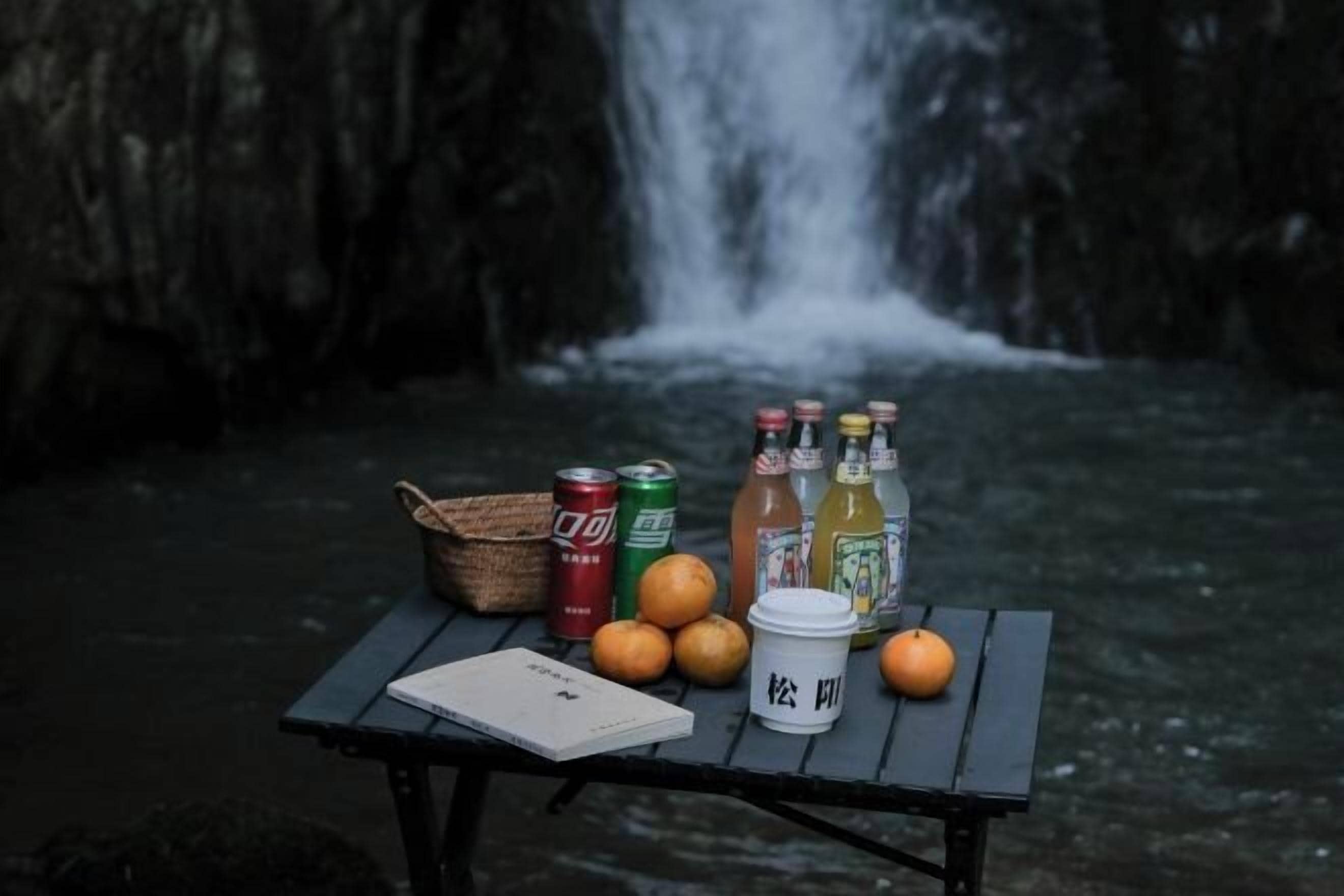 A table with beverages, a basket, and a notebook by a waterfall.