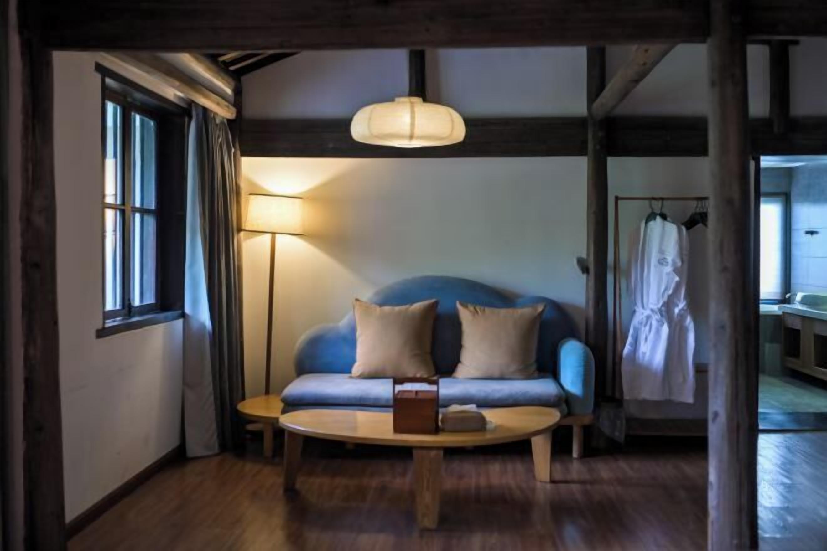 Een gezellige kamer met een blauwe bank, een houten bijzettafeltje, een hanglamp en een raam met gordijnen.