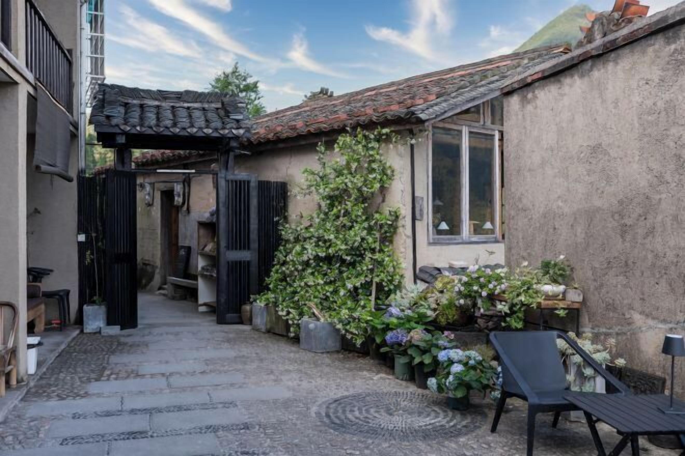 Une cour étroite surmontée d’un toit en tuiles, un petit jardin agrémenté de plantes en pot et un espace aménagé avec une chaise et une table.