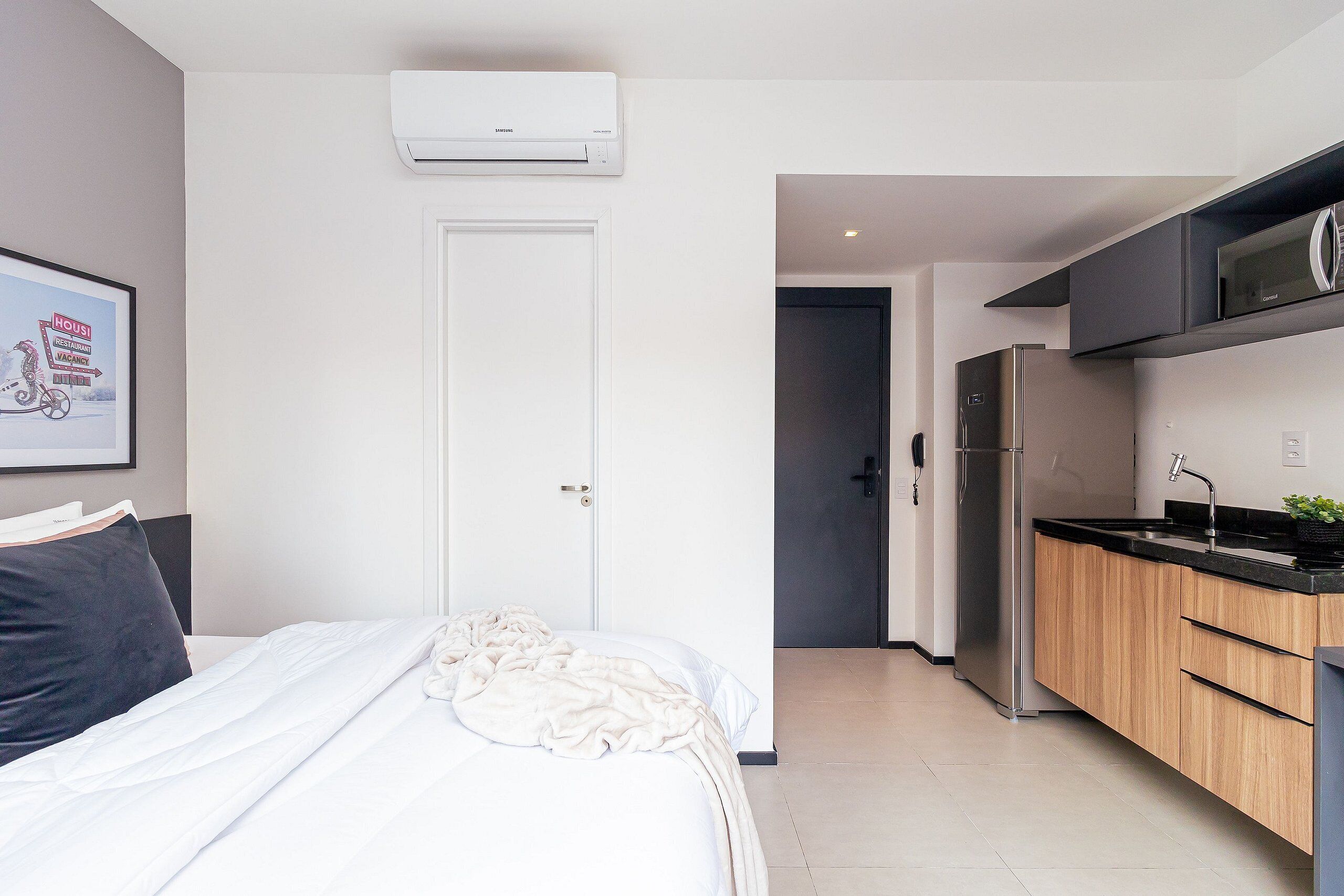 Een moderne hotelkamer met een bed, witte muren, een wandairconditioner, een keukenhoek met houten kasten en een roestvrijstalen koelkast.