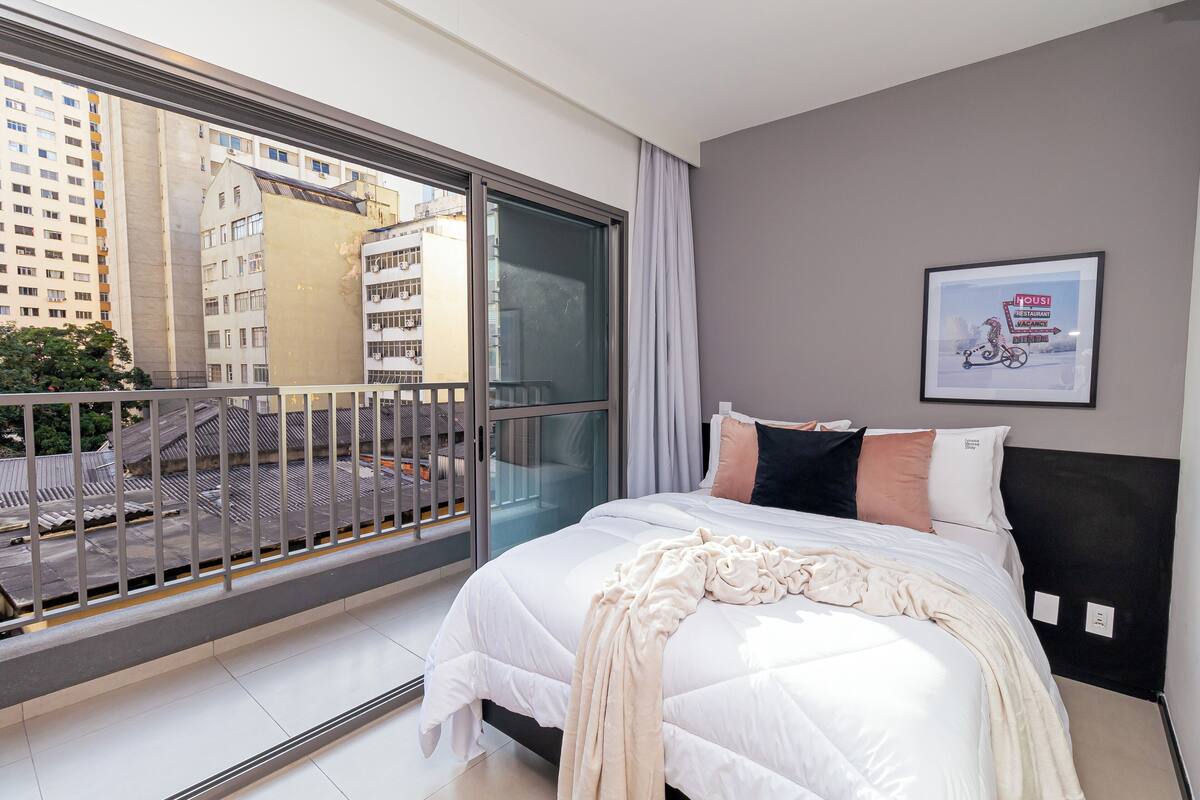 Quarto de hotel moderno com uma cama grande, varanda com vista para edifícios da cidade e um quadro na parede.