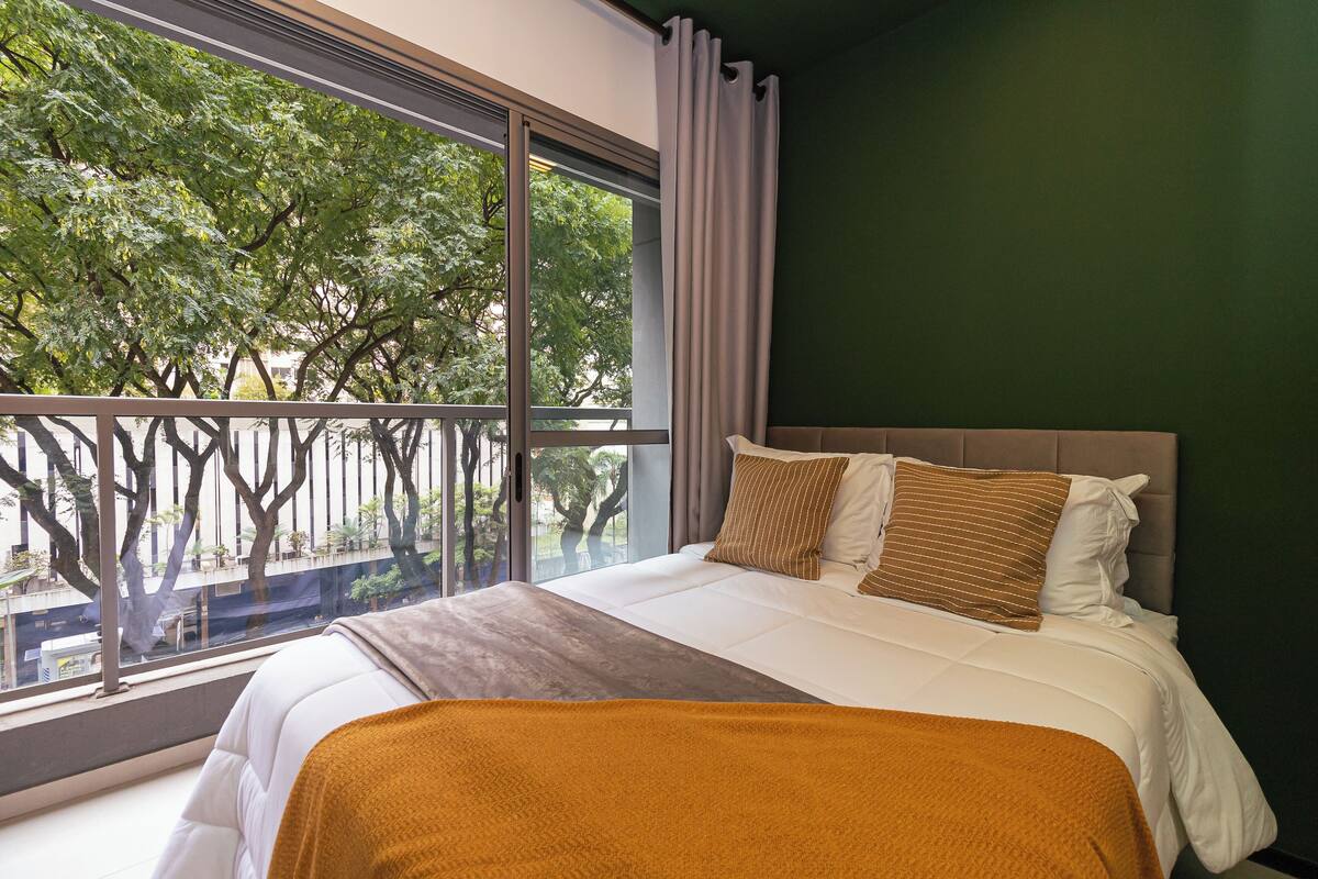 Quarto de hotel com uma janela grande, uma cama com roupa de cama branca e vista para árvores lá fora.