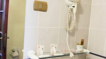 Un baño con inodoro blanco, un secador de pelo fijado en la pared, estante de vidrio con artículos de aseo y una toalla sobre el asiento del inodoro.