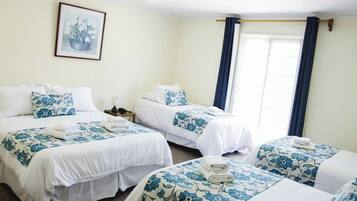 A hotel room with two beds, each with white bedding and blue floral patterns. There is a framed picture on the wall, a bedside table with a lamp, and a window with blue curtains.
