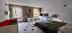 Une chambre comprenant un lit, un bureau, une chaise, un petit coin cuisine et une fenêtre donnant sur des immeubles.