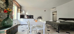 Une pièce compacte comprenant un coin cuisine, un espace repas avec une table et des chaises, un lit recouvert d’une couvre-lit vert foncé, et un grand vase vert orné de fleurs, posé sur une commode.