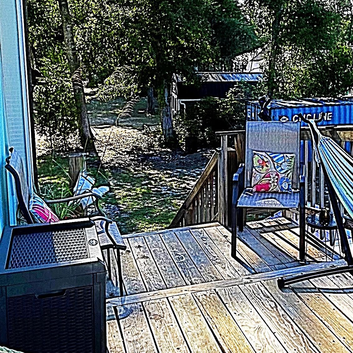 Une terrasse en bois offrant une vue sur une rivière, une chaise avec un coussin coloré et un panneau indiquant « PISTE CYCLABLE ».