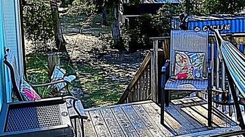 Une terrasse en bois offrant une vue sur une rivière, une chaise avec un coussin coloré et un panneau indiquant « PISTE CYCLABLE ».