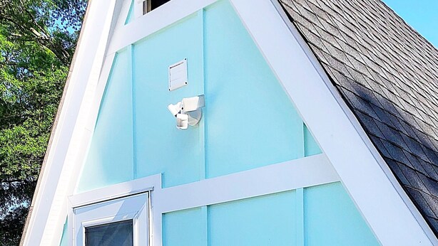Un edificio azul claro con puerta y ventana blancas, un respiradero pequeño y una terraza de madera con un tapete colorido.