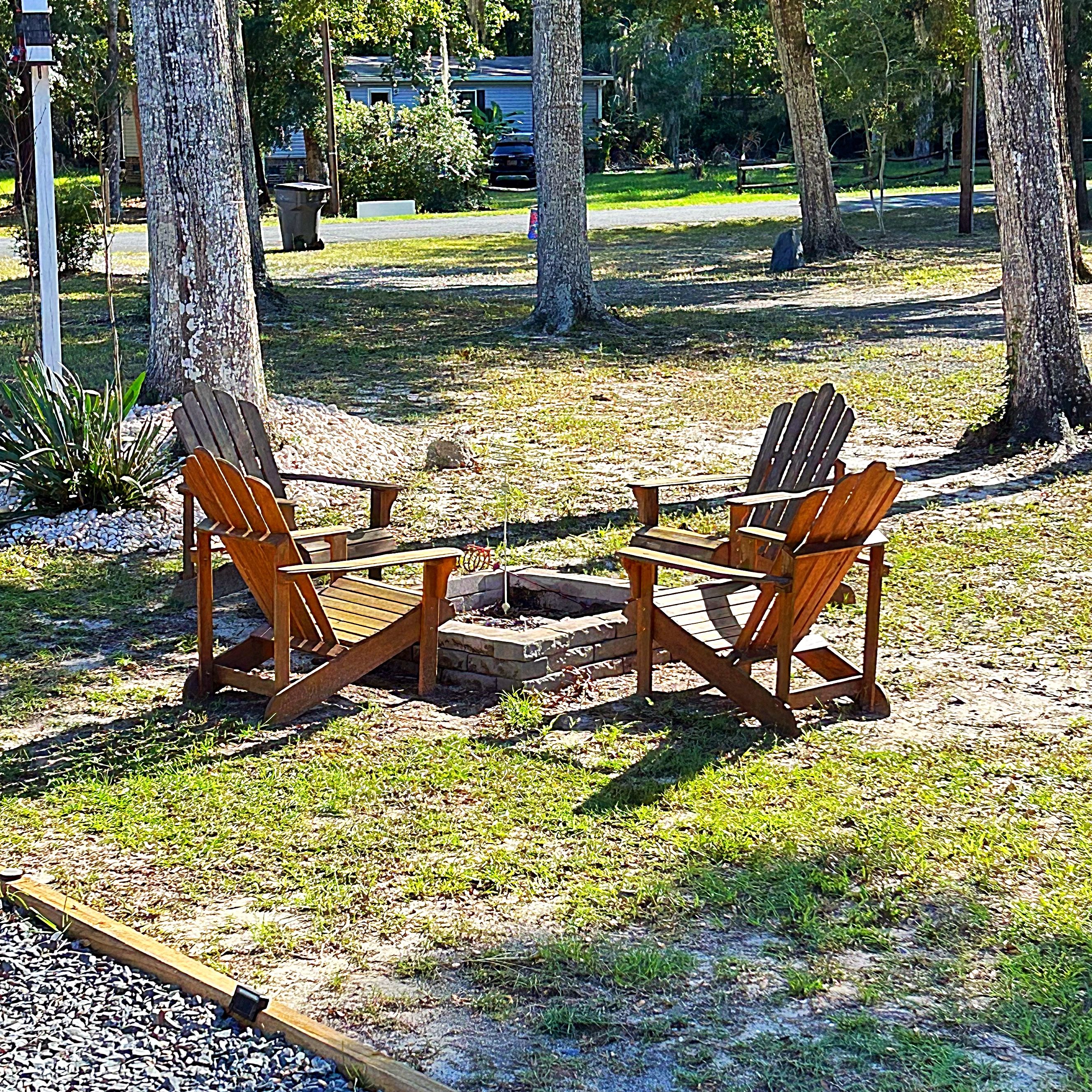 Two Adirondack chairs and a small table set up on a gravel area surrounded by trees.