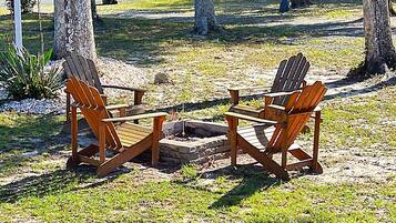 Two Adirondack chairs and a small table set up on a gravel area surrounded by trees.