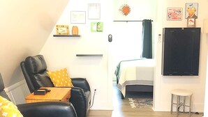 Una sala de estar moderna con un sofá de cuero negro, una mesa de centro de madera y una cama con cubrecama blanco y amarillo. Hay estantes en la pared con objetos decorativos y un televisor de pantalla plana.