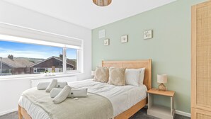 Een slaapkamer met een groot raam, een bed met een houten hoofdeinde, een nachtkastje met een lamp en een rieten kast.