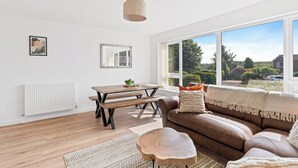 Een moderne woonkamer met houten vloer, een bruine leren bank, een houten salontafel, een eettafel met banken en een groot raam met uitzicht op groen en huizen.