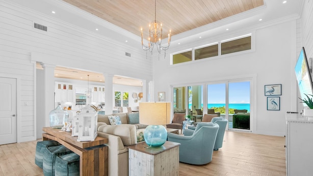 A spacious living room with a wooden ceiling, a chandelier, and a view of the ocean through large windows. - Yumey Sands Villa, Heated Pool & Unbelievable Year-Round Calm Beach (Moore Hill)