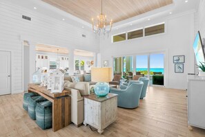 A spacious living room with a wooden ceiling, a chandelier, and a view of the ocean through large windows.