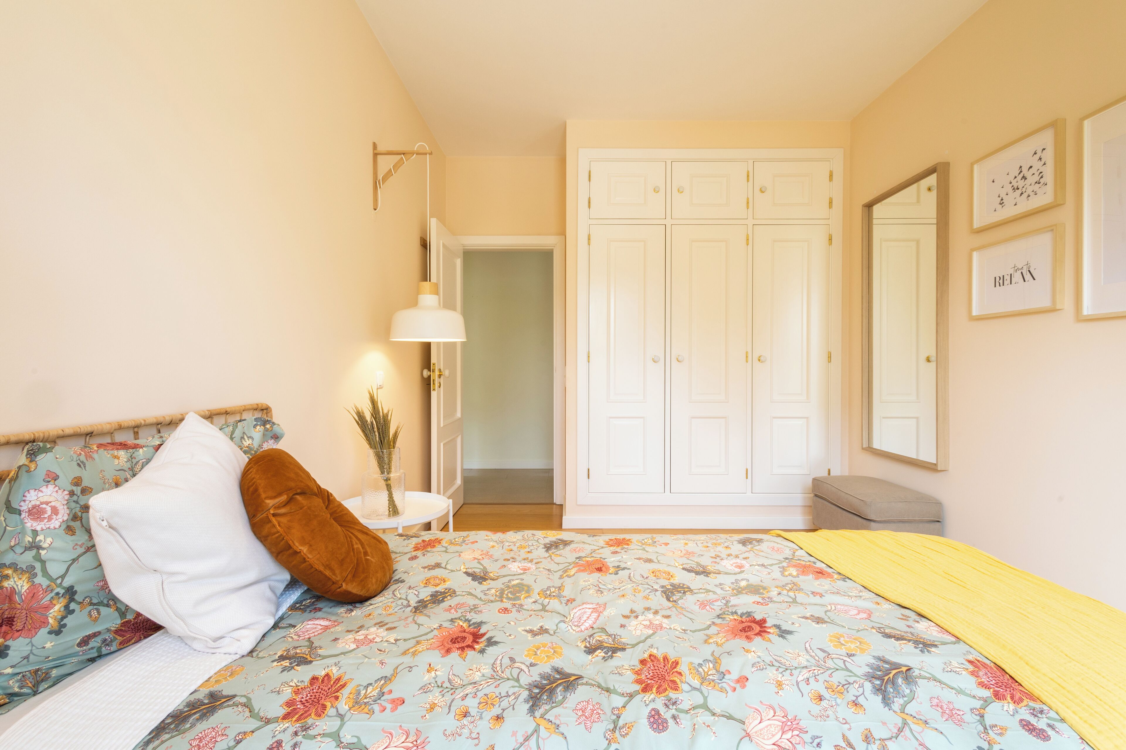 A bedroom with a floral bedspread, a bedside lamp, a wardrobe, and a mirror on the wall.