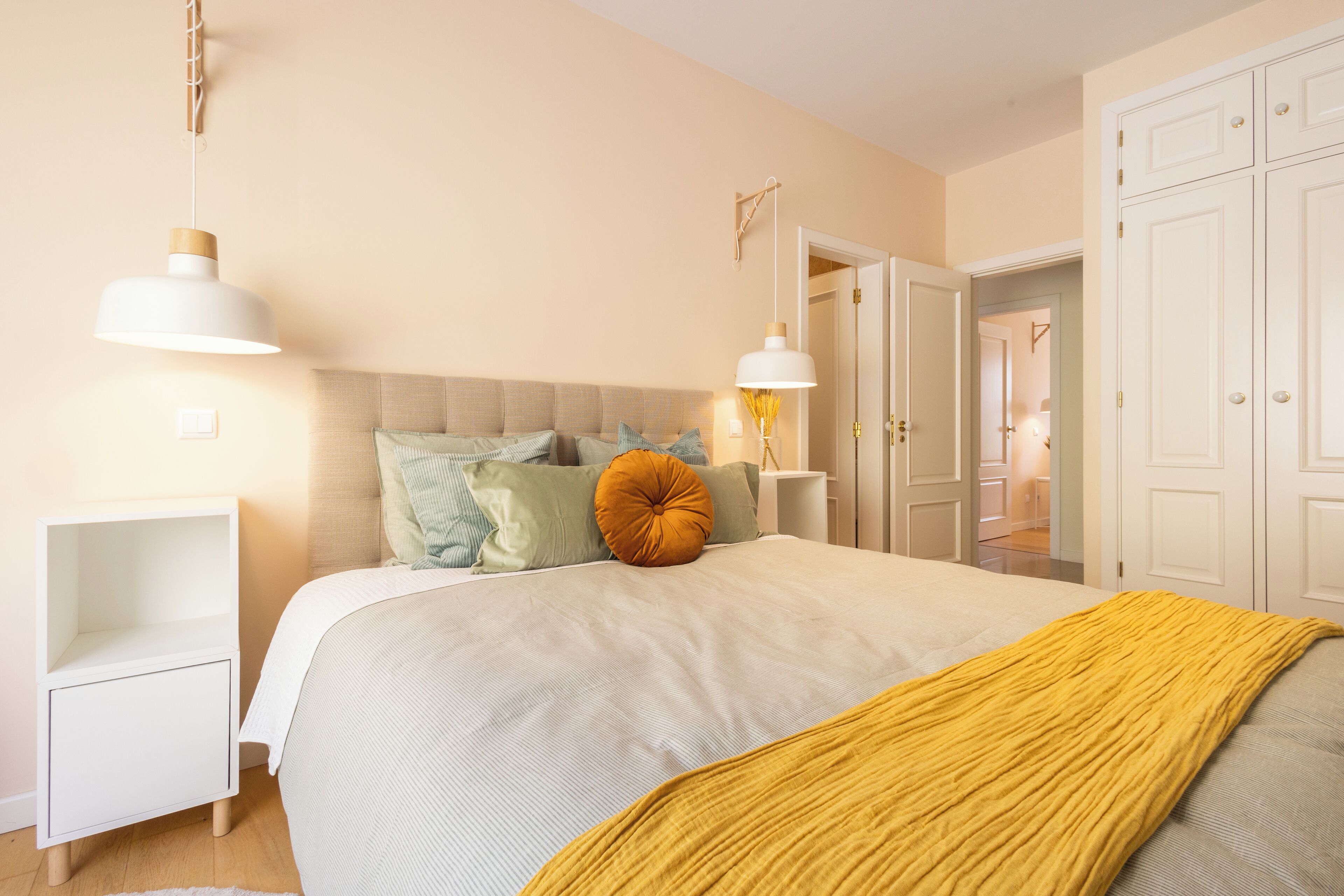 A bedroom with a bed, bedside tables, a nightstand, a wardrobe, and a door leading to another room.