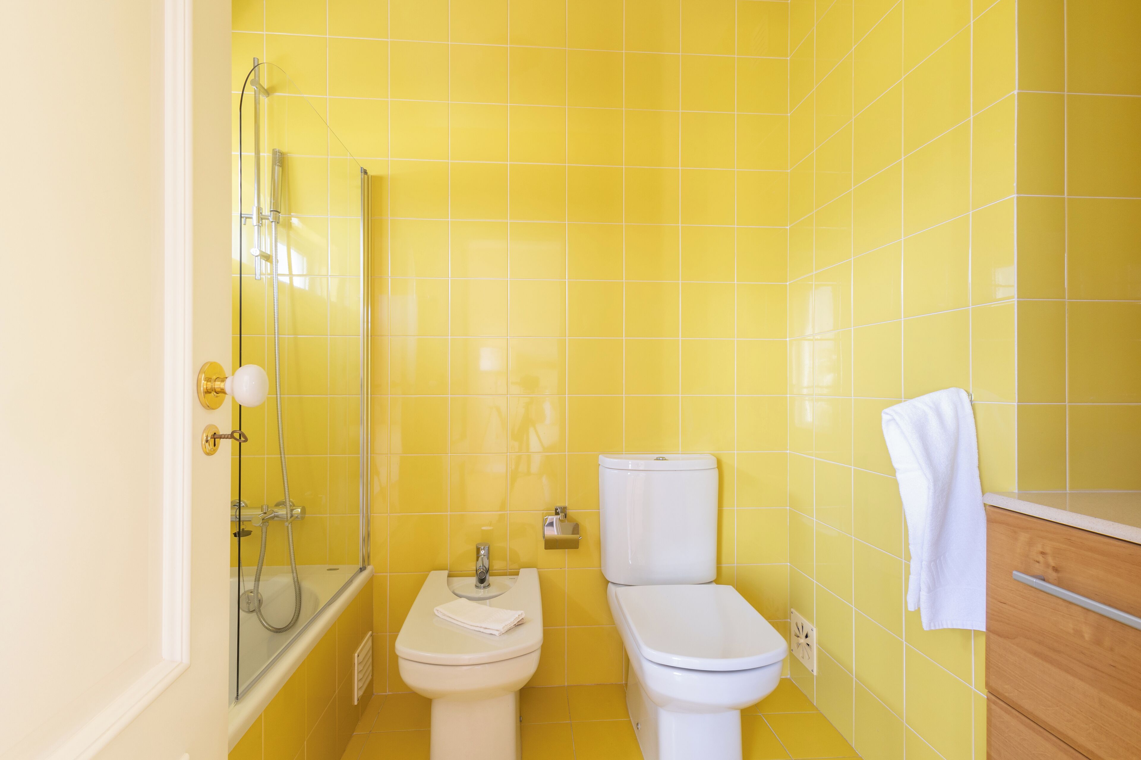 A bathroom with yellow tiles, a white toilet, a bidet, and a glass shower enclosure.