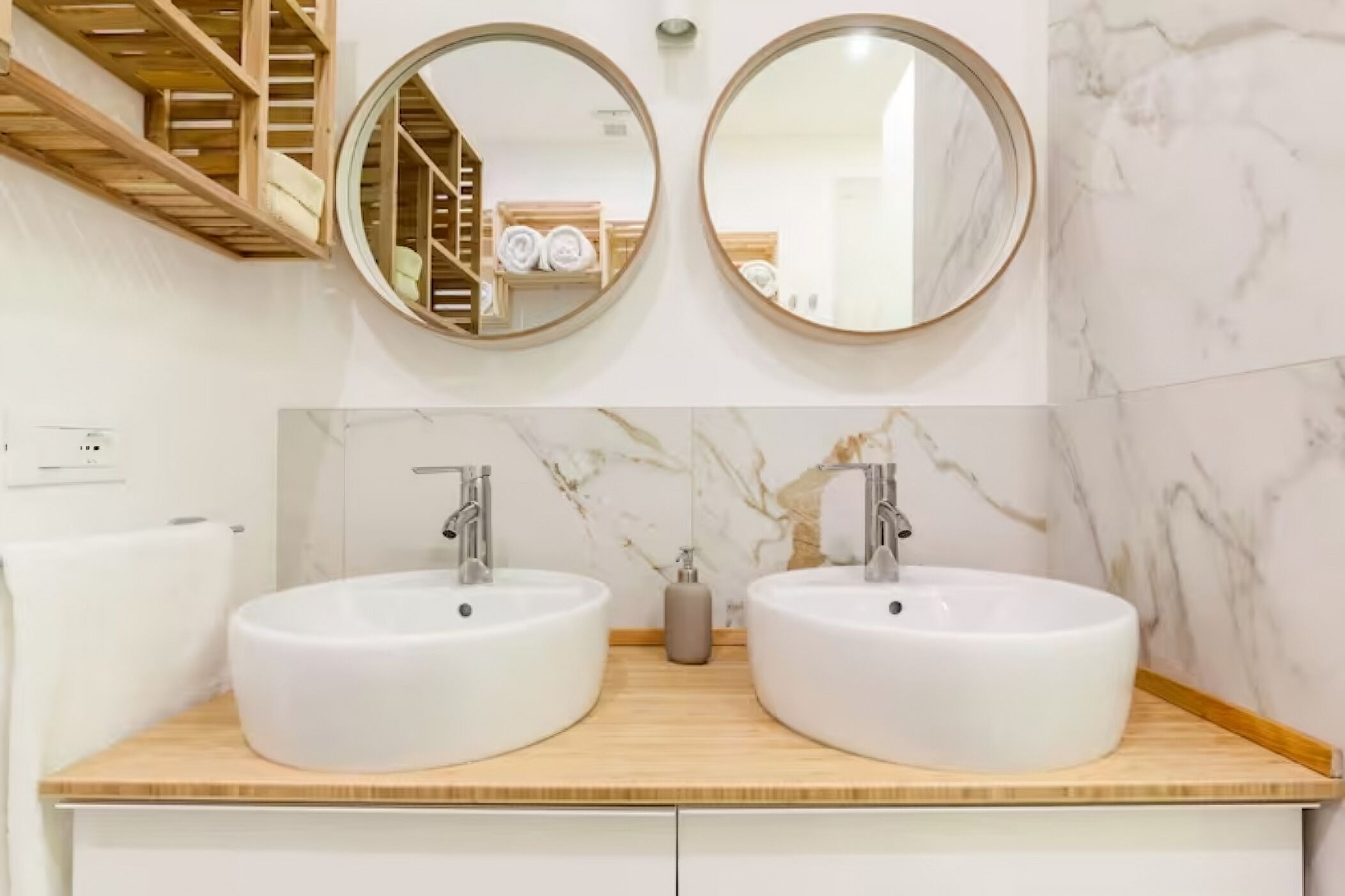 A bathroom with two round mirrors, a wooden shelf with towels, and a marble backsplash.