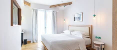 Un dormitorio con suelo de madera, una cama con ropa blanca, una mesita de noche, una lámpara de pared y una ventana con cortinas.