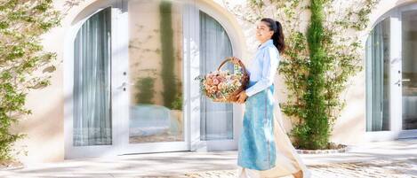 A person walking on a cobblestone path, carrying a basket of flowers, in front of a building with arched windows and greenery.