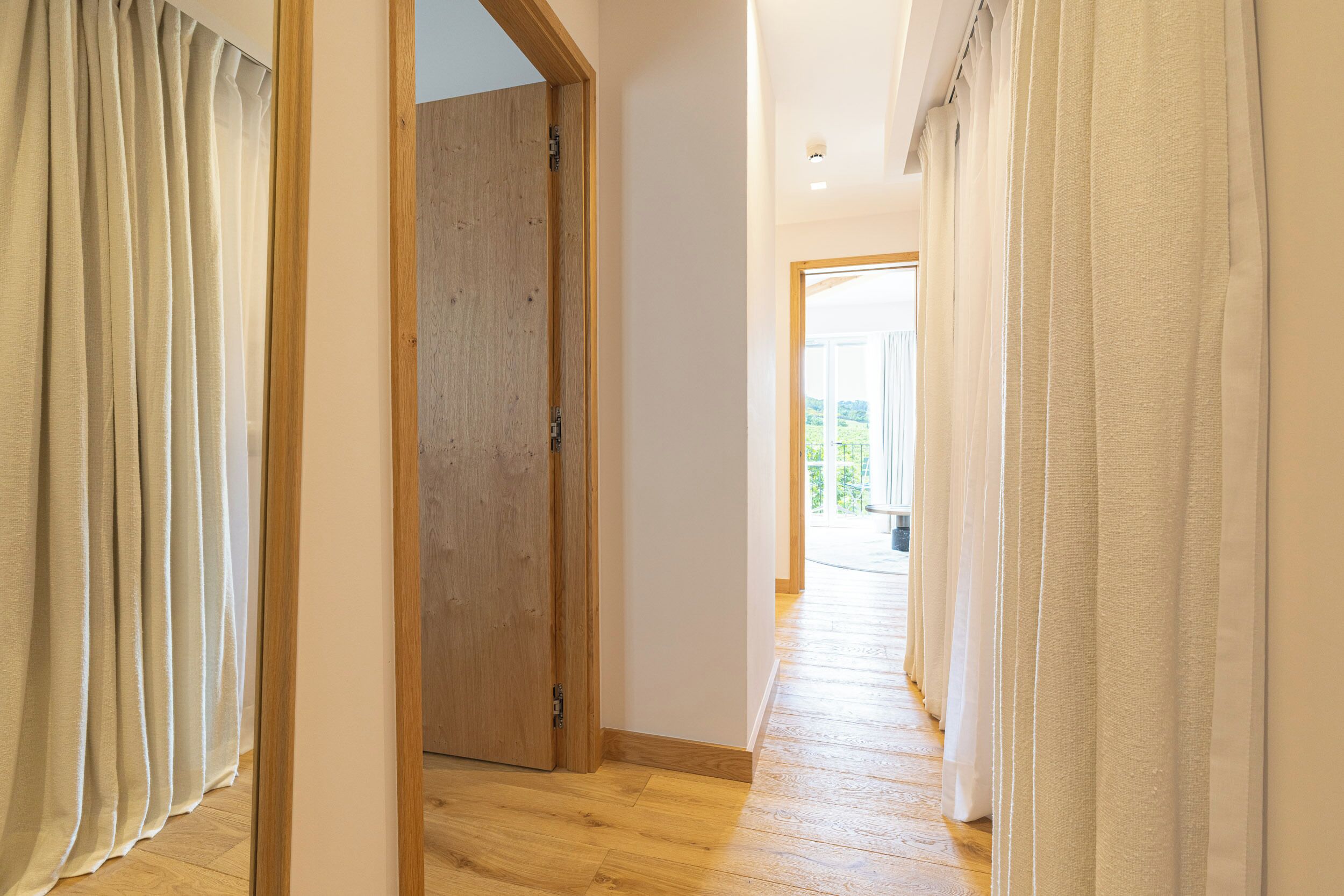 A hallway with wooden flooring, a wooden door, and white curtains.