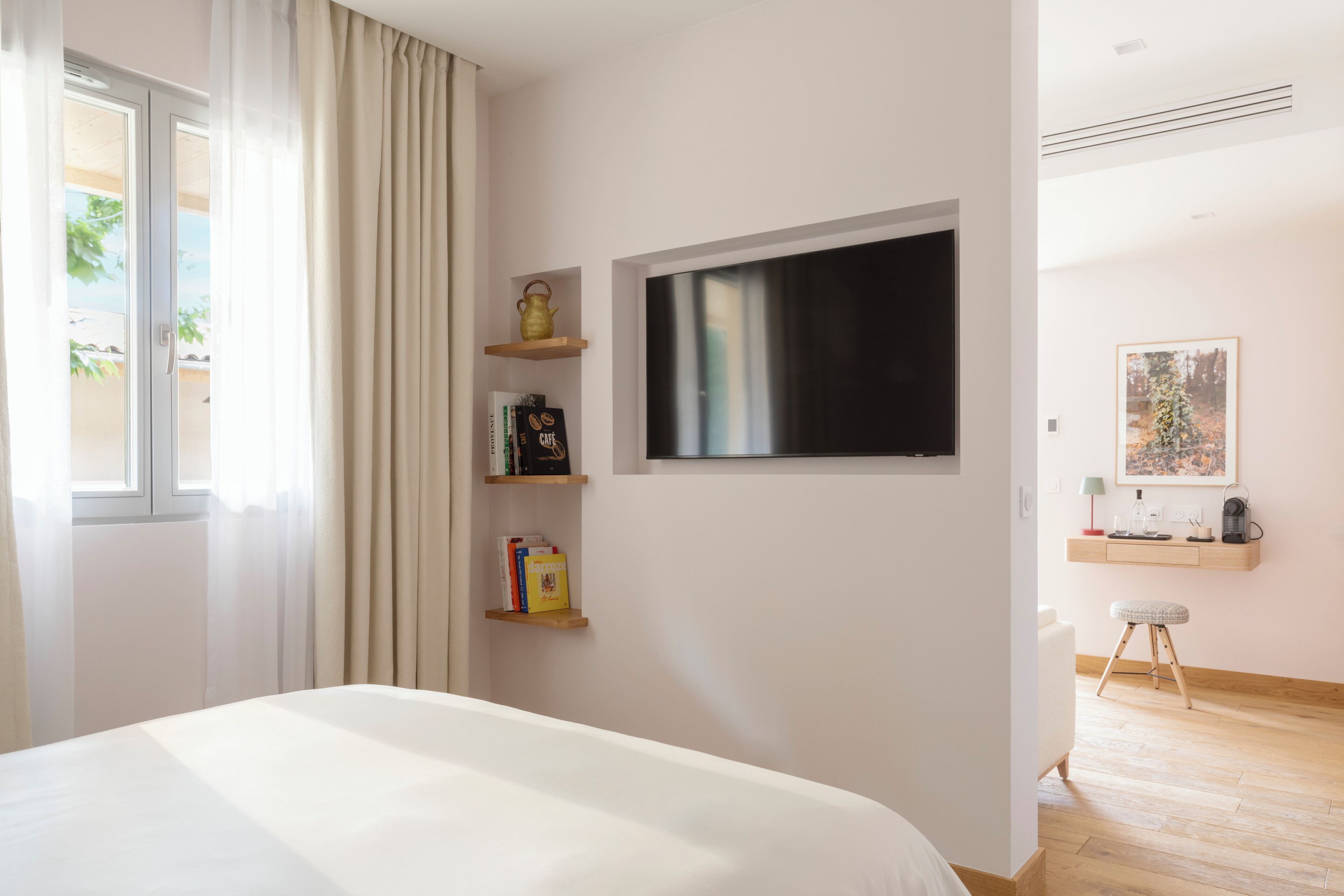 Quarto de hotel com cama, televisão na parede, pequena escrivaninha com cadeira e janela com cortinas.