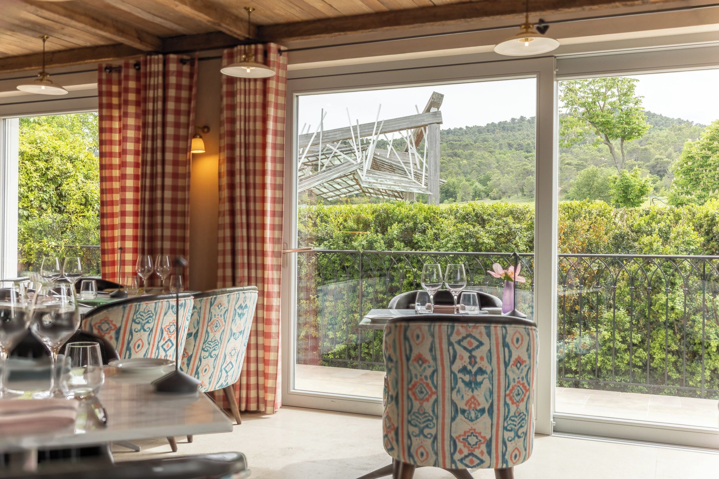 Un coin repas avec une table dressée pour deux personnes, une chaise à motifs et une vue sur un jardin luxuriant et des collines, le tout visible à travers de grandes fenêtres.