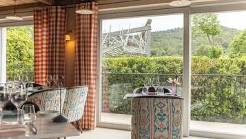 Un coin repas avec une table dressée pour deux personnes, une chaise à motifs et une vue sur un jardin luxuriant et des collines, le tout visible à travers de grandes fenêtres.