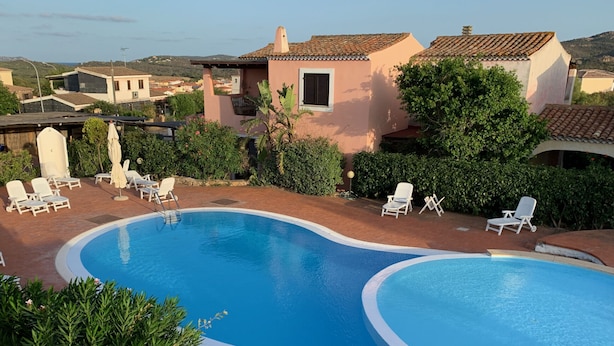 Piscina con sillas de descanso y una pérgola, rodeada de vegetación y edificios.
