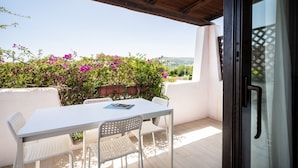 Un patio techado con una mesa y sillas blancas, un libro sobre la mesa, y vistas a un jardín con flores moradas.