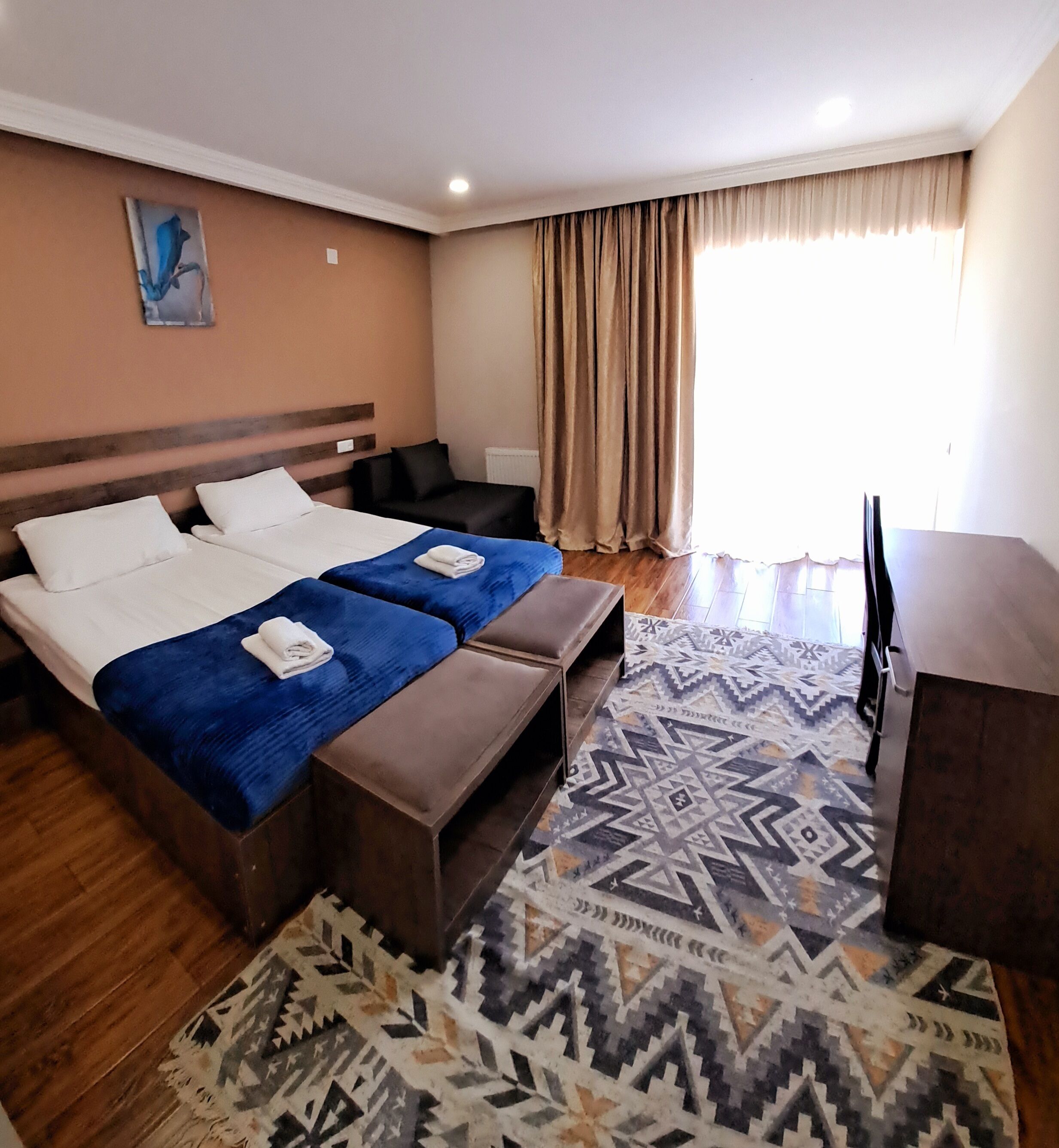 Habitación de hotel con cama, dos mesitas de noche, un escritorio, una silla, televisor y una alfombra estampada.