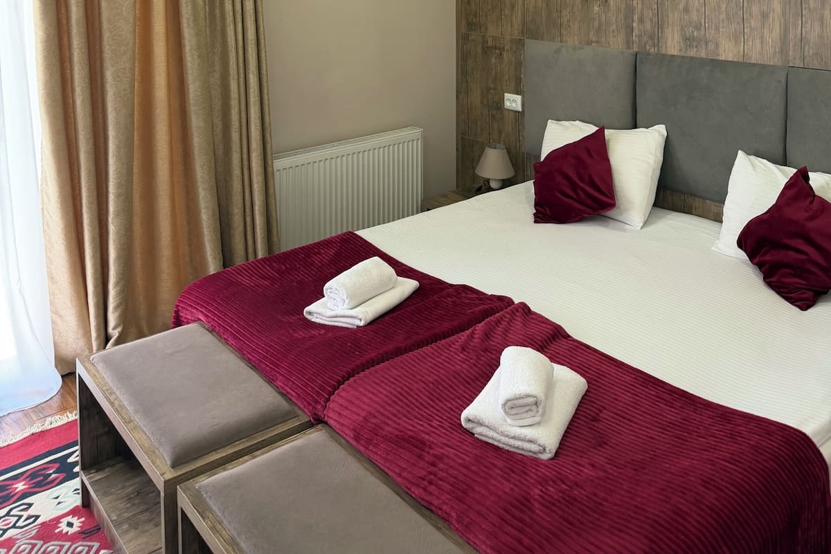 A hotel room with a large bed, a bench, a radiator, and a wooden panel wall.