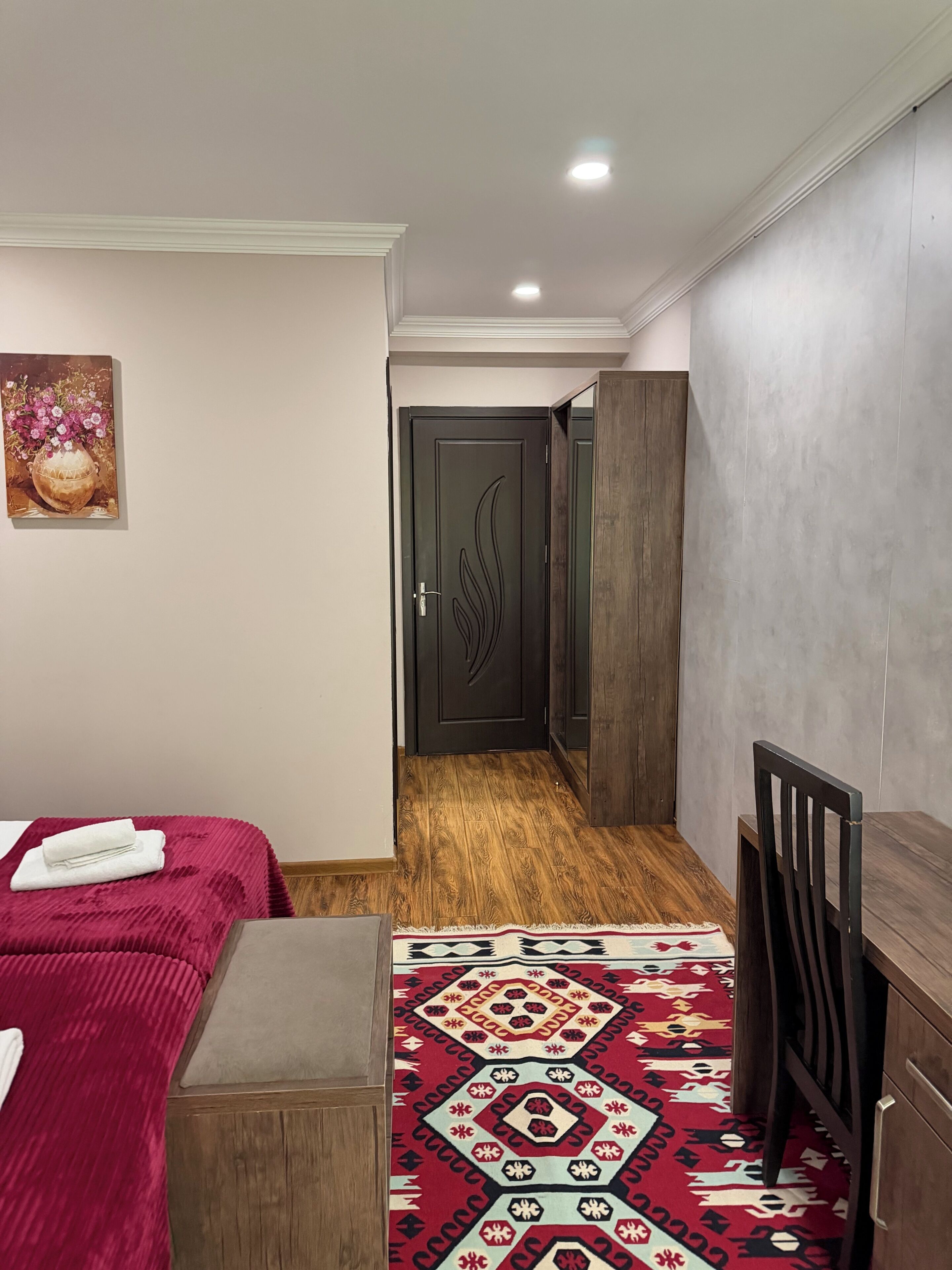 Habitación de hotel con una cama, una mesita, una silla, un tapete estampado, una puerta con diseño decorativo y un cuadro en la pared.