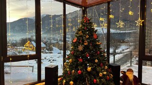 A Christmas tree decorated with ornaments and lights, a figure resembling Santa Claus, and a view of a snowy landscape through large windows. - Hotel Mountain Mestia (Mestia)