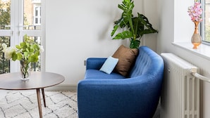 A room with a blue armchair, a round table, a potted plant, and a window with a view of buildings. - Lux Regents Park 2 bed 2 Bath Flat With AC (London)
