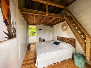 A bedroom with a wooden ceiling, a bed with a white bedspread, a colorful chair, and a small stool. - Sea Lodge : Maison Avec Vue Imprenable - Votre Refuge de Vacances Idéal (Sainte-Anne)
