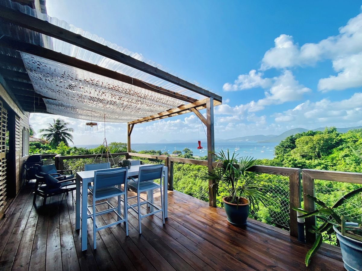 A wooden deck with a table and chairs, overlooking a lush landscape and the sea.