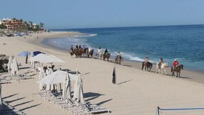 A beach with horse riders, beach umbrellas, and a building in the background. - Spend your Christmas week in Cabo at Villa La Valencia (San José del Cabo)