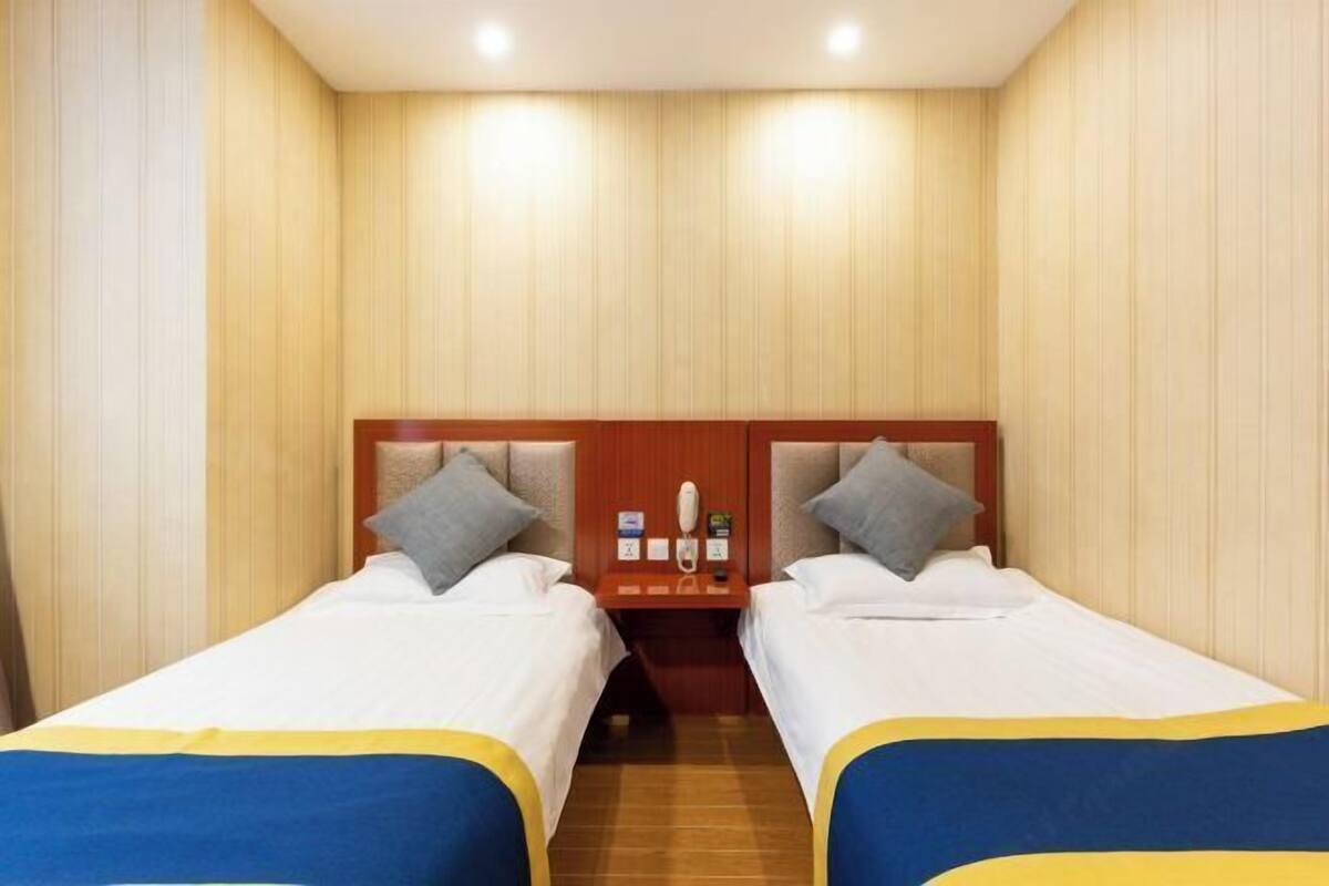 A hotel room with two beds, wooden headboards, a small bedside table with a phone, and a wall-mounted light fixture.